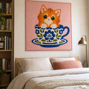 Peut inclure: Une tenture murale carrée présentant une illustration matelassée d'un chaton orange duveteux aux grands yeux verts assis dans une tasse à thé blanche ornée d'un motif floral bleu et vert, sur un fond rose clair uni.