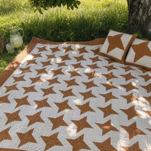 May include: A quilted blanket featuring a repeating pattern of brown stars on a white background with a brown border, accompanied by two matching square throw pillows, each displaying a single large brown star centered on a white field.