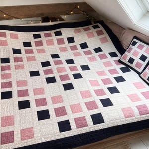 May include: A large square quilt and two matching throw pillows featuring a geometric grid pattern. The design consists of alternating solid black, light pink, and dusty rose squares separated by a cream-colored fabric border with visible stitching.