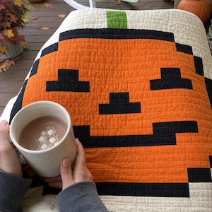 Puede incluir: Una manta acolchada con un diseño de calabaza naranja pixelada con ojos, nariz y boca negros, rematada con un tallo verde sobre un fondo crema, acompañada de una taza de cerámica moteada llena de chocolate caliente y malvaviscos.