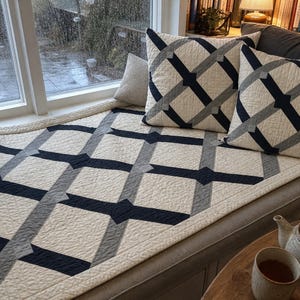May include: A quilted throw blanket and two matching square throw pillows featuring an off-white base with a geometric lattice pattern in dark navy blue and grey. The fabric has a visible quilted texture throughout the entire surface area.