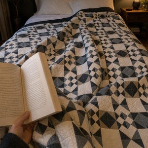Könnte beinhalten: Eine Steppdecke mit einem geometrischen Muster in Anthrazit, Hellgrau und Off-White. Das Textil zeigt ein sich wiederholendes Blockdesign aus Dreiecken und Quadraten mit sichtbaren Steppnähten auf der gesamten Stoffoberfläche.