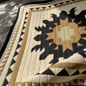 May include: A quilted blanket featuring a geometric star pattern in black, tan, and cream. The design includes a border of repeating black and tan triangles, with a grid-like stitched texture throughout the fabric surface and a folded corner.