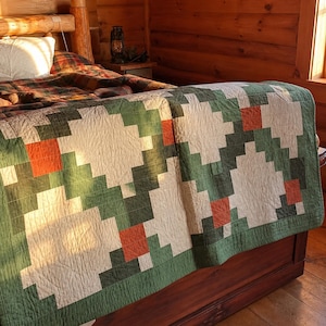 Könnte beinhalten: Ein rechteckiger Steppdecke mit einem geometrischen Muster aus weißen, dunkelgrünen und rostorangen Quadraten. Der Stoff weist eine sichtbare gitterartige Steppstruktur auf der gesamten Oberfläche auf, die ein sich wiederholendes Blockdesign erzeugt.