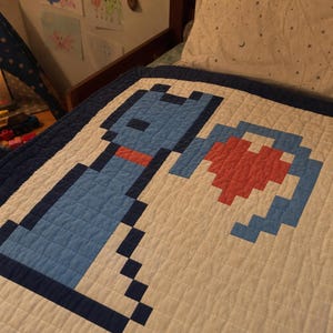 Peut inclure: Une courtepointe blanche présentant un motif de chat bleu pixélisé avec un collier rouge et un cœur rouge. La courtepointe possède une bordure bleu foncé et est confectionnée avec des motifs de surpiqûres carrées visibles sur toute la surface.