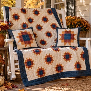 May include: A quilted throw blanket and two matching square throw pillows featuring a repeating star pattern in navy blue, burnt orange, and cream. The textile set has a cream-colored base with a dark navy blue border and visible grid stitching.