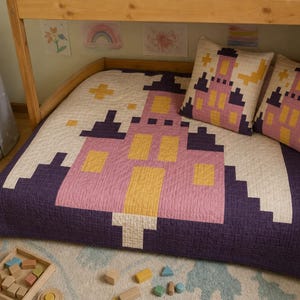 May include: A quilted bedspread and two matching square pillows featuring a pixelated castle design in shades of purple, pink, and yellow on a cream background. The bedding set is displayed on a wooden bed frame with a light-colored rug underneath.