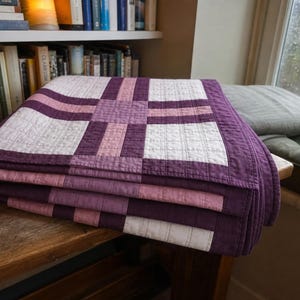 May include: A folded quilt featuring a geometric pattern in shades of deep purple, light purple, and off-white. The fabric displays a visible grid of straight-line quilting stitches throughout the surface, creating a textured appearance across the fabric.