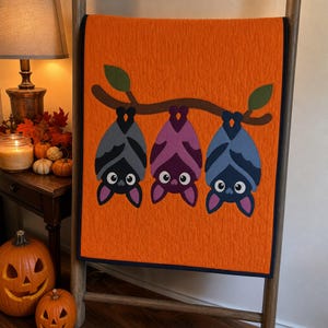 Op de afbeelding: Een feloranje quilt met een applicatieontwerp van drie cartoonvleermuizen die ondersteboven aan een bruine boomtak met twee groene bladeren hangen. De vleermuizen zijn uitgevoerd in tinten grijs, paars en blauw met donkere omlijningen.