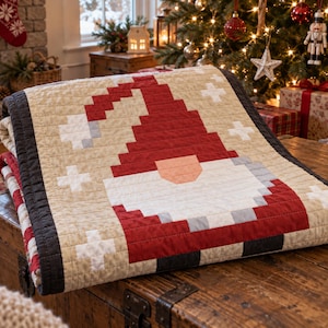 May include: A quilted throw blanket featuring a pixelated gnome design in red, white, gray, and beige. The gnome wears a tall red hat and has a large white beard. The quilt is finished with a dark charcoal gray border and white cross accents.
