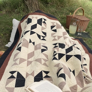 May include: A quilt featuring a geometric pattern of cream, charcoal black, and muted mauve triangles and squares. The textile has a wide border in dark charcoal and a thin inner border in rust brown, with visible quilted stitching throughout the fabric.