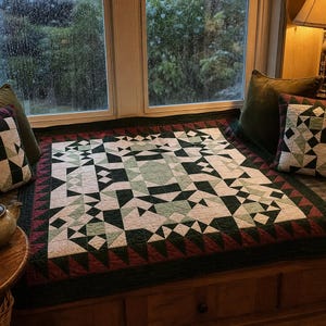 May include: A square quilt featuring a geometric pattern in shades of dark green, light green, cream, and maroon. The quilt has a central star-like design surrounded by a border of small triangles in dark green and maroon, with matching pillows.