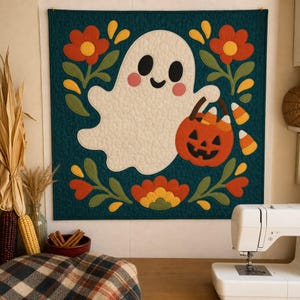 May include: A square quilted wall hanging featuring a central white ghost with black eyes and pink cheeks, holding an orange jack-o'-lantern bucket filled with candy corn. The design is set against a dark teal background with red flowers.