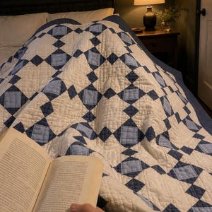 May include: A quilt featuring a geometric pattern of cream, light blue, and dark blue fabric squares and diamonds. The textile displays a repeating grid design with visible stitching throughout, covering a bed surface in a dimly lit room.
