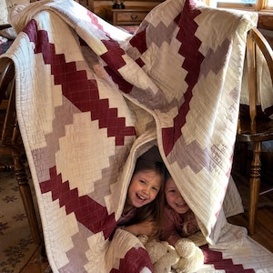 May include: A quilt featuring a geometric pattern in cream, muted mauve, and deep burgundy. The fabric has a visible grid-like stitched texture throughout. The quilt is draped over chairs, creating a tent-like structure with visible seams.