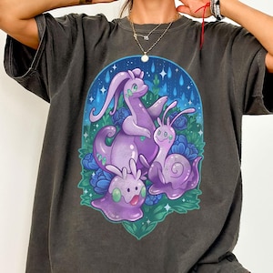 Peut inclure: Un t-shirt à manches courtes gris anthracite avec une illustration graphique centrale représentant trois créatures violettes ressemblant à des escargots et des limaces, entourées de fleurs bleues et de gouttes de pluie sur fond bleu.