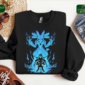 Puede incluir: Una sudadera negra de manga larga con un estampado gráfico de una criatura azul brillante grande detrás de una silueta oscura más pequeña de la misma criatura, rodeadas por llamas de energía azul y efectos de luz.