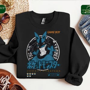 May include: A black long-sleeve sweatshirt featuring a graphic of the blue and black Lucario character. The design includes the text GAME BOY, POCKET MONSTERS in Japanese and English, and small icons of Poké Balls and handheld gaming consoles.