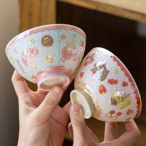 以下が含まれることがあります： 表面に質感のある2つのセラミック製ボウル。左側のボウルは水色でピンクのアクセントがあり、茶色のビスケット、黄色い星、ピンクの形のイラストが描かれています。右側のボウルは薄いピンク色で赤のアクセントがあり、イチゴ、サクランボ、黄色い猫のイラストが描かれています。