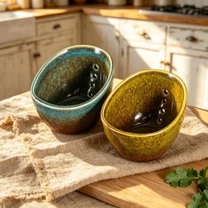 May include: Two ceramic bowls with an angled rim and a series of small drainage holes along the interior side. One bowl features a deep teal and brown reactive glaze, while the other displays a vibrant olive green and brown mottled finish.