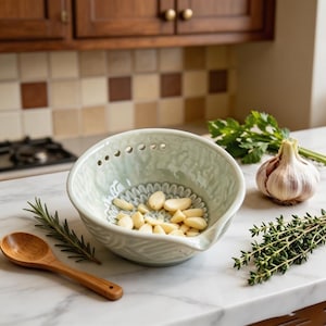 May include: A light green ceramic bowl with a textured interior and a row of small holes near the rim, containing several peeled garlic cloves. A wooden spoon, a sprig of rosemary, a head of garlic, and a sprig of thyme are placed nearby.