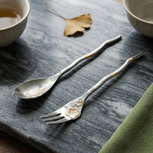 May include: A silver-toned metal spoon and fork set featuring slender, wavy handles and gold-coloured floral accents on the bowl of the spoon and the base of the fork tines. Each piece includes a small gold-coloured detail along the handle.