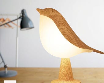Op de afbeelding: Een vogelvormige tafellamp met een lichtgekleurde houtnerfafwerking op de kop, staart en voet, met een gloeiend, doorschijnend wit lichaam dat zacht licht uitstraalt. De lamp staat op een ronde voet op een houten ondergrond.