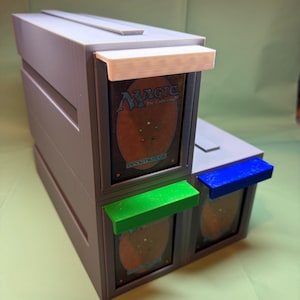 May include: A grey 3D-printed storage box for trading cards with three vertical compartments. Each compartment features a protruding handle in white, green, or blue, and displays a Magic: The Gathering card back with the text Deckmaster visible.