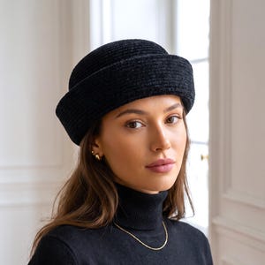 May include: A black textured cloche hat with a rounded crown and a structured, upturned brim. The hat features a dense, slightly fuzzy fabric construction with a consistent dark tone, providing a clean and uniform appearance for this headwear piece.