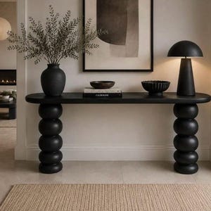 Peut inclure: Une console noire avec deux pieds formés de sphères empilées, surmontée d'un vase noir contenant des branches vertes, une pile de livres, un petit bol noir et une lampe de table noire en forme de champignon. Le meuble a un plateau allongé lisse.