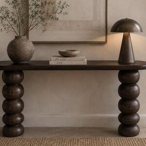 May include: A dark brown wooden console table featuring an elongated oval top supported by two vertical columns composed of stacked spherical shapes. The table surface holds a round textured vase with branches, a small bowl, and two stacked books.