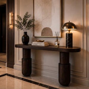 Peut inclure: Une console en bois brun foncé avec deux pieds cylindriques, surmontée d'un vase noir contenant du feuillage vert, deux livres empilés étiquetés Chanel, un petit diffuseur en verre et une lampe en marbre noir avec un abat-jour dôme doré.