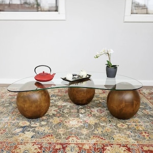 May include: A coffee table featuring a clear, organic-shaped glass top supported by three large, spherical wooden bases with a medium-brown finish. On the glass surface sits a red cast-iron teapot, a small rectangular tray, and a white orchid in a pot.