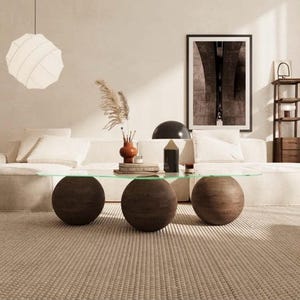 May include: A coffee table featuring a clear glass top supported by three large, dark brown wooden spheres. The table is positioned in front of a light-colored sofa, with a textured rug underneath and a decorative vase and lamp on the surface.