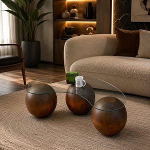 Puede incluir: Una mesa de centro con un tablero de cristal transparente y curvado, sostenido por tres grandes bases esféricas de madera en color marrón oscuro. La mesa sostiene una pequeña taza de cerámica blanca y un objeto decorativo verde cuadrado.