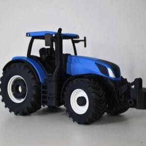 Op de afbeelding: Een blauwe speelgoedtractor met een zwarte uitlaat, zwarte banden met diep profiel en witte velgen. Het voertuig is voorzien van een zwart frontgewicht, een cabine met een zwarte zitting en zijspiegels aan de buitenkant.