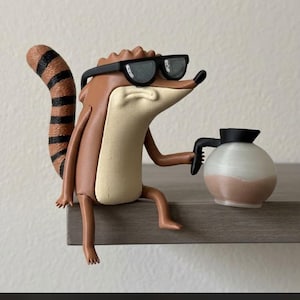 Puede incluir: Una figura de mapache de color marrón y crema con gafas de sol negras, cola a rayas y expresión gruñona. La figura sostiene en su mano derecha una pequeña cafetera redonda de color blanco y marrón con asa y tapa negras.