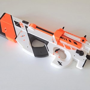 May include: A white, orange, and black toy blaster with a futuristic bullpup design. The body features geometric patterns, black accents, and small graphic symbols including an X inside a diamond shape. It has a top rail and a short barrel.
