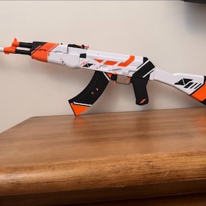 Puede incluir: Un rifle de juguete con un esquema de colores blanco, negro y naranja brillante. El rifle tiene un cañón largo, un cargador curvo y una culata con acentos geométricos negros y naranjas. El diseño incluye pequeños símbolos blancos.