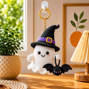 May include: A crochet ghost keyring featuring a white ghost body with black eyes and pink cheeks, wearing a black witch hat with a purple band and yellow buckle. The ghost holds a small black crochet bat with white fangs and pink cheeks.