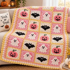 May include: A crochet blanket featuring a grid of square motifs in pink, white, and yellow. The squares display alternating patterns of pink jack-o'-lanterns, white ghosts with pink bows, and black bats, all crafted with yarn in a granny square style.