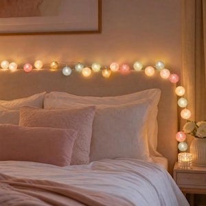 Op de afbeelding: Een decoratieve lichtsnoer met bolvormige lampenkappen in diverse pastel- en neutrale kleuren. De lampjes zijn verlicht, stralen een warme gloed uit en zijn horizontaal over een hoofdeinde en verticaal langs de zijkant gedrapeerd.
