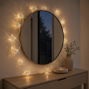 Peut inclure: Un miroir rond avec un cadre sombre fin est entouré d'une guirlande de lumières sphériques en métal perforé. Les lumières projettent une lueur chaude et à motifs sur le mur, créant une bordure décorative autour de la surface en verre circulaire.