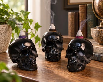 May include: Three black skull-shaped incense burners made of a glossy material. Each skull features a conical incense cone placed on top, emitting thin wisps of white smoke that curl upward into the air, creating a distinct visual effect.