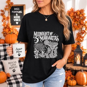 May include: A black short-sleeve t-shirt featuring a white graphic print with the text Midnight Margaritas, a margarita glass, a coffee maker, and stars. A white mug with the text Aussie Mom and a paw print icon is held in one hand.