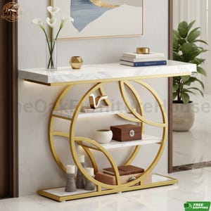 May include: A console table featuring a white faux marble top and base with a gold-toned metal frame designed in a circular geometric pattern. The table includes two interior shelves holding decorative items, including a vase, books, and boxes.