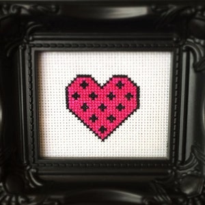 Puede incluir: Una imagen enmarcada en negro con un fondo blanco y un corazón rosa con lunares negros en el centro. El marco tiene un diseño decorativo y el texto "Sugar Stitch" es visible en la esquina inferior derecha.