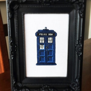 May include: A framed cross-stitch of a blue police box with the text "Police Box" on the side. The cross-stitch is in a black frame with ornate detailing.