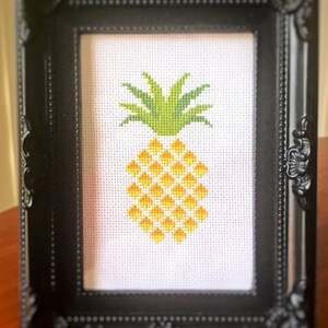 May include: A framed cross-stitch of a yellow pineapple with green leaves. The frame is black with ornate detailing. The image is on a wooden table.
