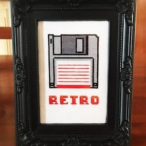 May include: A framed cross-stitch design featuring a pixelated floppy disk with the word "RETRO" underneath. The frame is black with ornate detailing.